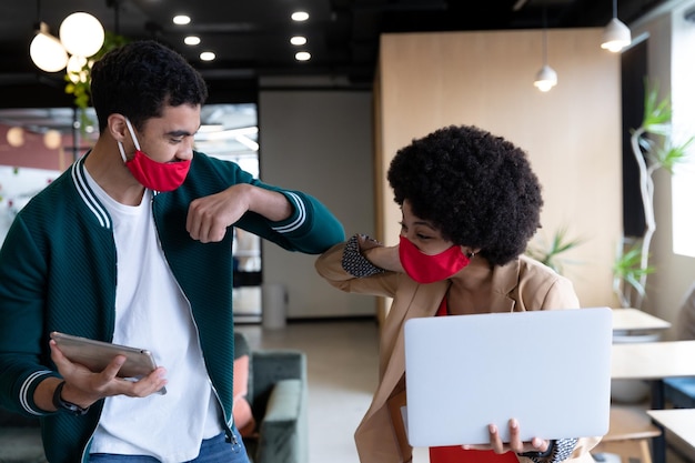 Diverse businessman and businesswoman wearing face masks touching elbows in office. social distancing in business office workplace during covid 19 coronavirus pandemic.