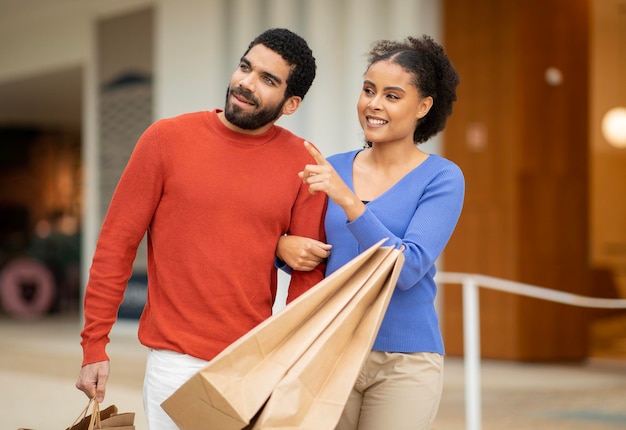 Diverse couple shopping pointing finger aside holding bags at mall