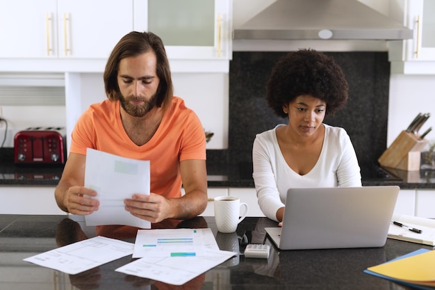 Diverse couple sitting in kitchen using laptop and paying bills