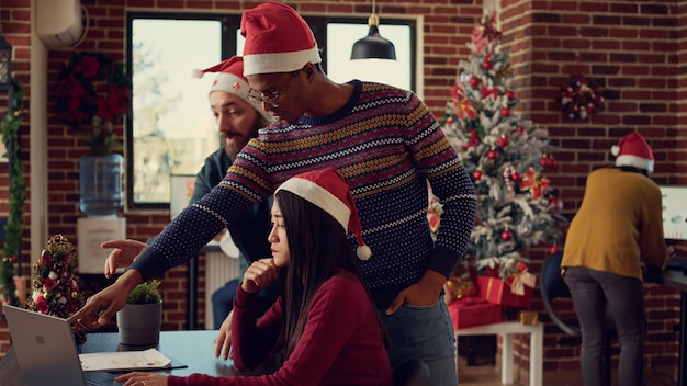 Photo diverse coworkers working on startup report in festive office, doing teamwork at desk during christmas time. brainstorming ideas on computer in space decorated with holiday ornaments.