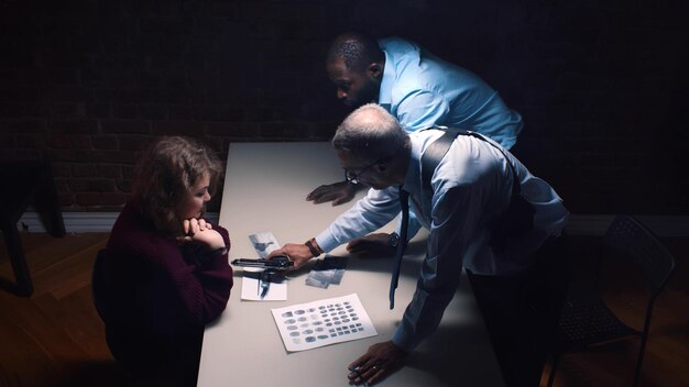 Photo diverse detective interrogating scared arrested woman and threatening with gun