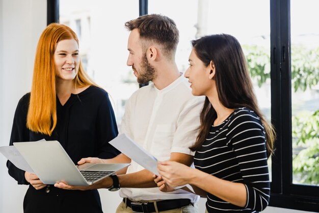 Diverse ecstatic businesspeople discussing papers and working on laptops at an office table while smiling