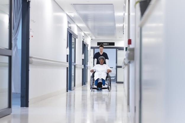 Diverse female doctor walking with senior female patient in wheelchair in hospital corridor