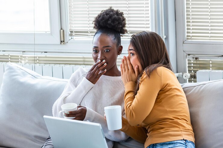 Photo diverse female friends sharing secrets two women gossiping at home excited emotional girl whispering to her friend ear while sitting in livingroom