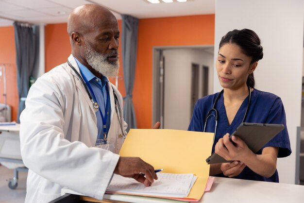 Diverse female and senior male doctor using tablet and talking in hospital ward, copy space. Hospital, medical and healthcare services.