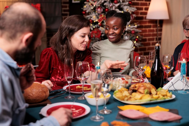 クリスマスのホーム パーティーでおしゃべりをする多様な友人、お祝いのディナー テーブルで話す女性、伝統的なクリスマス フードを食べる人々。美しい豪華な装飾が施された場所での冬の休日のお祝い