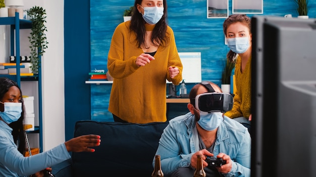 Diverse friends trying to win playing video games experiencing
virtual reality headset having fun enjoying beer and popcorn
wearing face mask to prevent spreading virus in time of global
outbreak