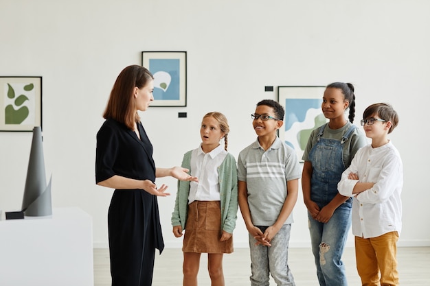 Diverse groep kinderen die naar vrouwelijke experts luisteren tijdens een bezoek aan een moderne kunstgalerie, kopieer ruimte