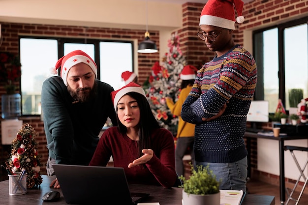 Diverse groep mensen die werken tijdens kerstavond, teamwork doen om een zakelijk project op kantoor te plannen met kerstverlichting en feestelijke decoraties. Met behulp van laptop op het werk, seizoensgebonden wintervakantie.