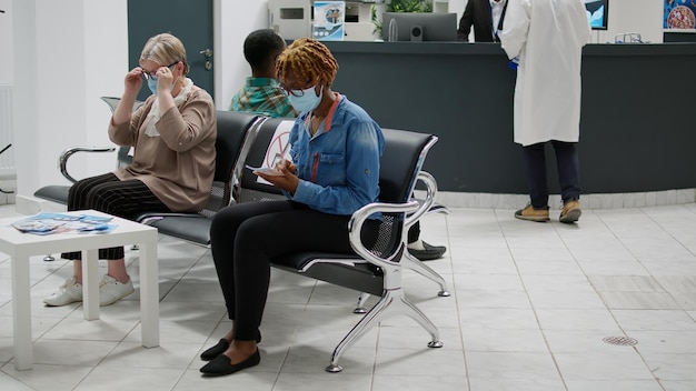 Diverse groep patiënten met gezichtsmaskers die in de wachtruimte bij de receptie van het ziekenhuis zitten en zich voorbereiden op het starten van een medische controleafspraak tijdens de covid 19-pandemie. Gezondheidszorg examen.