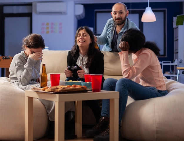 Diverse groep vrienden hecht zich tijdens het spelen van videogames op tv-console en verliest na het werk met joystickcontroller. multi-etnisch team geniet van een feest op kantoor met hapjes en drankjes