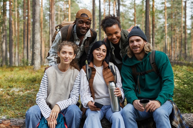 Diverse group of friends enjoying hike in forest and looking at camera while sitting on log copy space