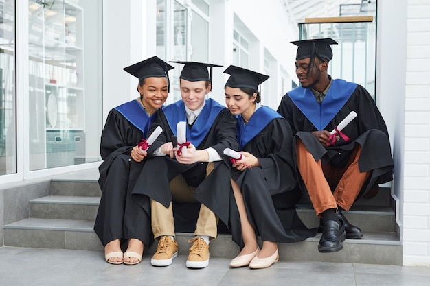 Diverse group of happy young people wearing graduation gowns and using smartphone while sitting on steps outdoors