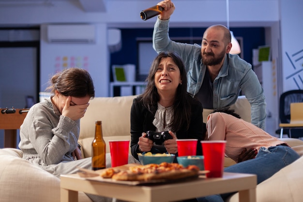 Diverse group of mates bonding while playing video game on tv\
console losing with joystick controller after work. multi ethnic\
team enjoy office celebration party with snacks and drinks