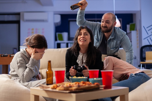 Diverse group of mates bonding while playing video game on tv console losing with joystick controller after work. Multi ethnic team enjoy office celebration party with snacks and drinks