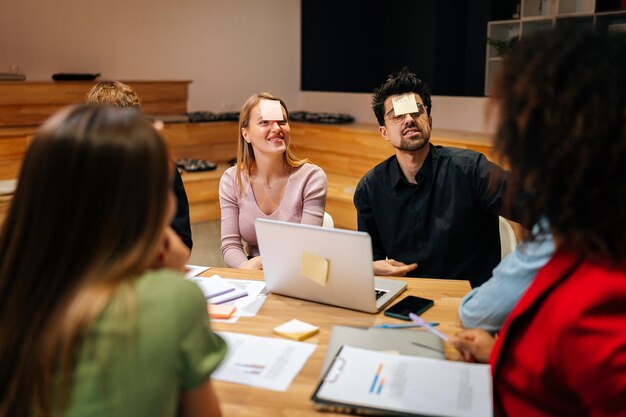 Diverse happy coworkers playing guessing game with sticky notes on forehead after work for entertainment Cheerful multiracial group of colleagues having fun