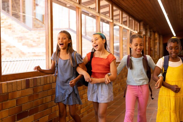 Photo diverse happy schoolgirls with bags walking and embracing in elementary school corridor