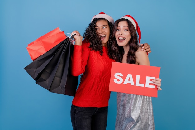 Diverse happy women with colorful shopping bags and red copyspace sale sign isolated over blue