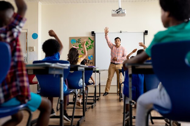 Diverse male teacher and elementary schoolchildren raising hands in class