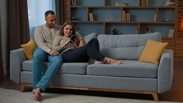 Diverse man and woman interracial couple homeowners relax on sofa talk video call communicate online