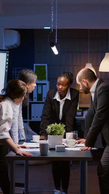 Diverse multi ethnic businesspeople discussing over graphs paperwork standing at conference table