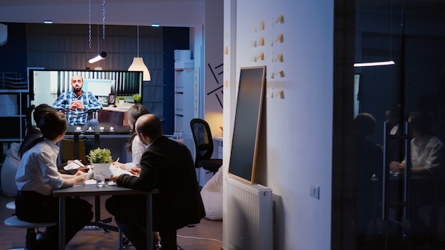 Diverse multi-ethnic businesspeople discussing with remote manager in wheelchair during online videocall conference meeting late at night in company office room. Focused teamwork working overtime