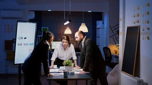 Diverse multi ethnic teamwork standing around table brainstorming company ideas checking management statistics paperwork. Focused coworkers planning overtime business presentation in meeting room