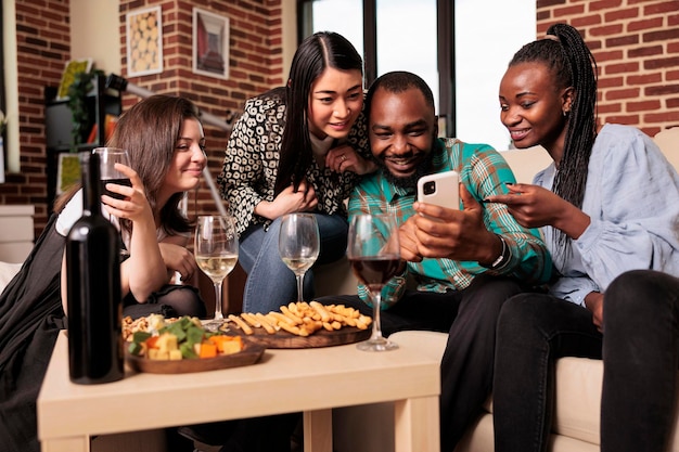 Diverse nationaliteitsgroep mensen staren naar smartphonescherm, glimlachen, lachen, genieten van vriendschap, alcohol drinken, wijn, kaas proeven, broodstengels op een zelfgemaakt feest.