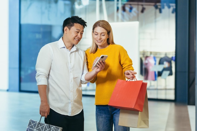 Diverse paar aziatische man en blonde vrouw in kleding supermarkt kijken naar het scherm van de mobiele telefoon