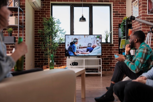 Diverse people watching soap opera tv show on channel,
gathering at home with beer and snacks to have fun. young people
looking at drama movie or film on television program,
relaxing.