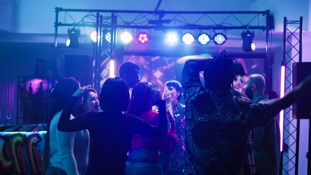 Diverse persons dancing on music, partying with crowd of people and having fun at nightclub with dj. group of friends enjoying disco party on dance floor at event. handheld shot