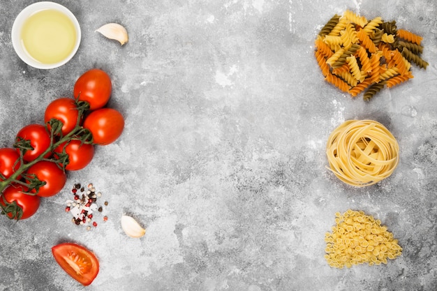 Diverse rauwe pasta en ingrediënten voor het koken (tomaten, olijfolie, knoflook) op grijze achtergrond. Bovenaanzicht, kopieer ruimte. Voedsel achtergrond