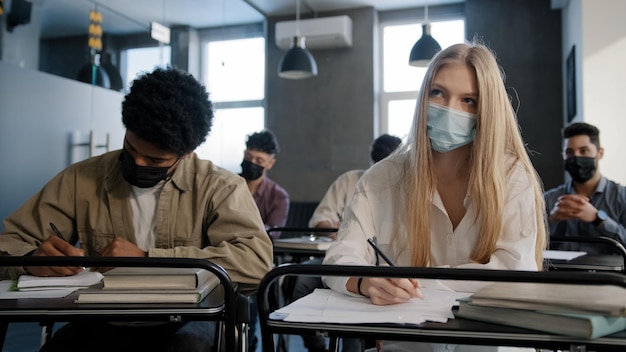 Diverse students in medical mask in classroom at lesson listen to lecture writing pupils studying