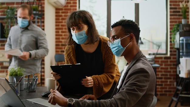 Photo diverse team of colleagues working on company data in office during covid 19 pandemic. using laptop to plan business charts presentation with documents, teamwork collaboration.