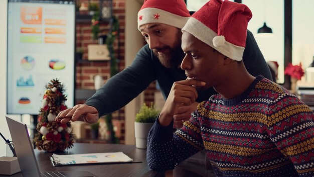 クリスマスの飾りで飾られたお祭りのオフィスでノートパソコンと書類を使用する男性の多様なチーム。クリスマスイブのお祝い、季節の装飾の間に職場でチームワークをしている人々。