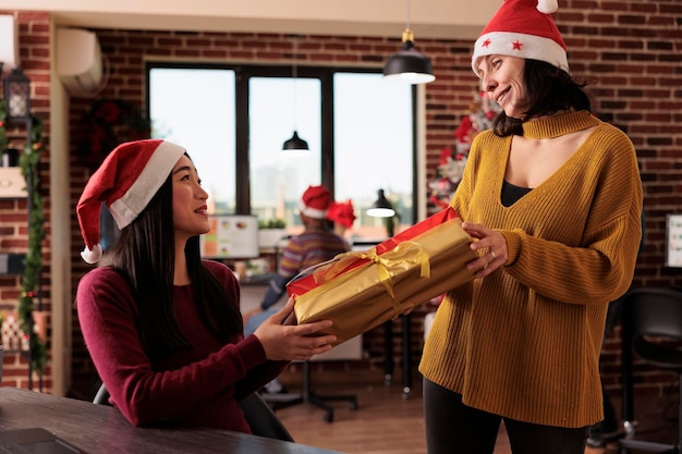 Diverse vrouwen wisselen de huidige doos uit op kerstavondviering op kantoor, in een ruimte versierd met kerstboom, lichten en decoraties. feestelijke mensen die geschenken geven tijdens de feestdagen.