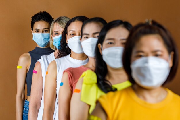 Diversiteitsgroep van vrouwen van multinationale etnische patiënten draagt gezichtsmasker in de rij staan op volgorde van hoogte, kijk naar de camera met kleurrijke pleisters op de schouder samen na vaccinatie.