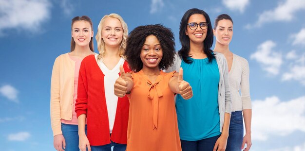 diversity, race, ethnicity and people concept - international group of happy smiling different women over white showing thumbs up over blue sky and clouds background