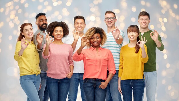 diversity, race, ethnicity and people concept - international group of happy smiling men and women showing ok hand sign over holidays lights background