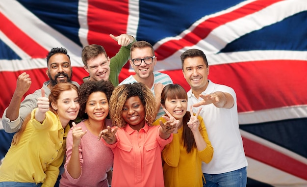 Разнообразие, раса, этническая принадлежность и концепция людей - международная группа счастливых улыбающихся мужчин и женщин, показывающих большой палец вверх и мир на фоне британского или английского флага