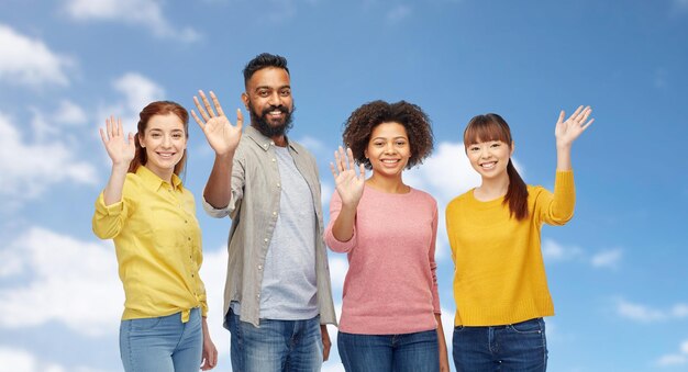 Photo diversity, race, ethnicity and people concept - international group of happy smiling men and women waving hands over blue sky and clouds background
