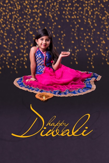 Diwali Greeting Card showing Cute little indian or asian girl in traditional wear holding a diya or Terracotta oil lamp. Front view over black background