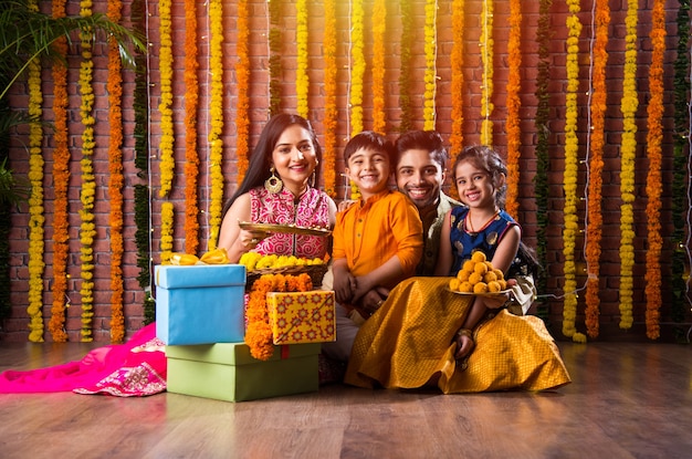 ディワリ祭またはラクシャバンダン祭-甘いラドゥー、石油ランプまたはディヤとギフトボックスでディーパバリまたはバイドゥージ祭を祝う4人のインドの若い家族、食べ物を食べる、または自撮りする