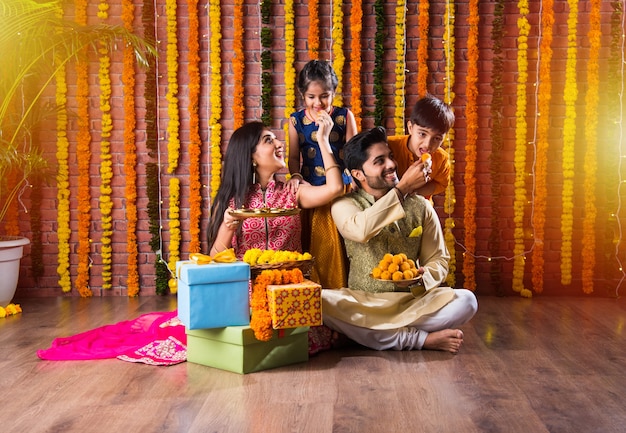 Diwali or Rakshabandhan Celebration - Indian young family of four celebrating Deepavali or bhai Dooj festival with sweet laddoo, oil lamp or diya and gift boxes, eating food or taking selfie