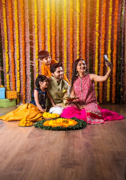 Diwali or Rakshabandhan Celebration - Indian young family of four celebrating Deepavali or bhai Dooj festival with sweet laddoo, oil lamp or diya and gift boxes, eating food or taking selfie