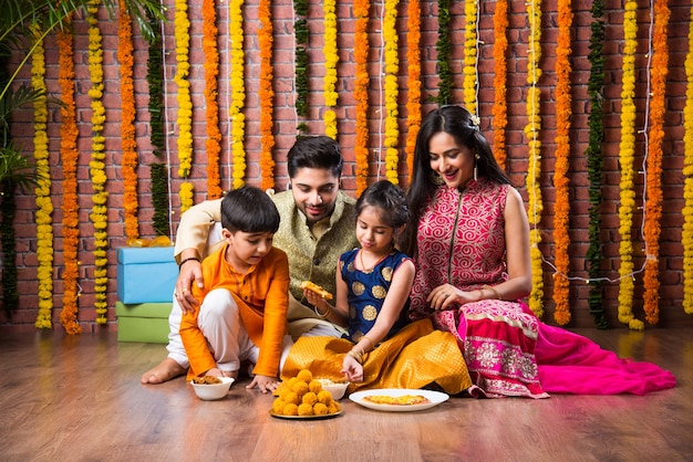 Diwali or Rakshabandhan Celebration - Indian young family of four celebrating Deepavali or bhai Dooj festival with sweet laddoo, oil lamp or diya and gift boxes, eating food or taking selfie