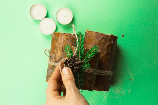 Diy christmas candle holder made of pine logs, candles, craft\
rope, fir branches and cones. hands attach the decoration.\
step-by-step instructions flat lay, step 4.