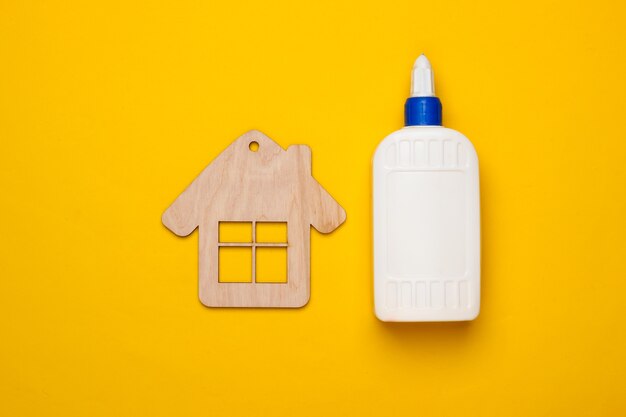 DIY house concept. Wooden mini house figurine and a bottle of glue on a yellow background. Top view