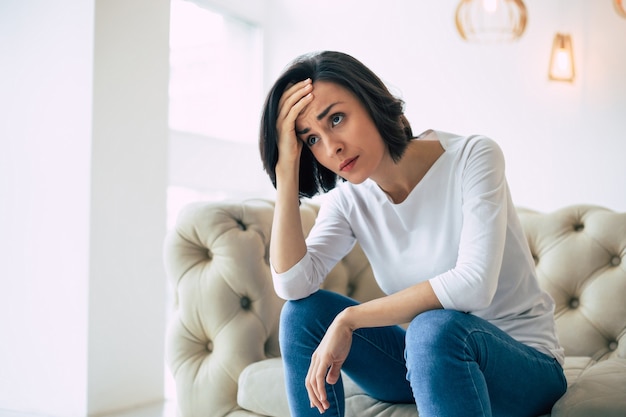 Dizziness. Side-view photo of a woman who is sitting on a couch and holding her forehead, while suffering from vertigo.