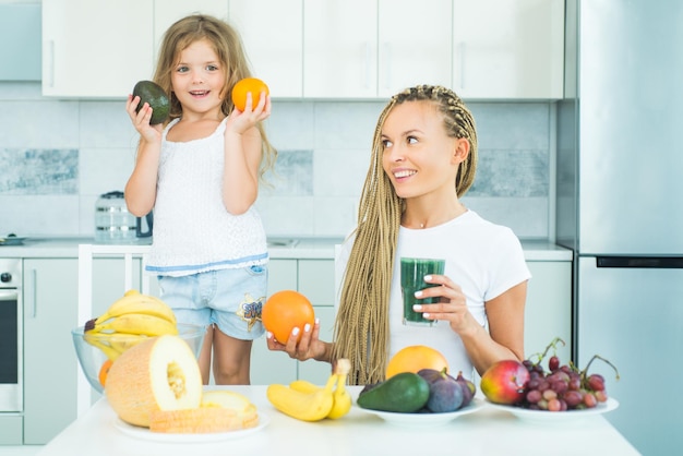 Dochter en moeder drinken groene smoothie en eten fruit Veganistische maaltijd en detox-menu Moeder en dochter drinken groene smoothie in de keuken Biologisch voedsel en vitamine voor familie